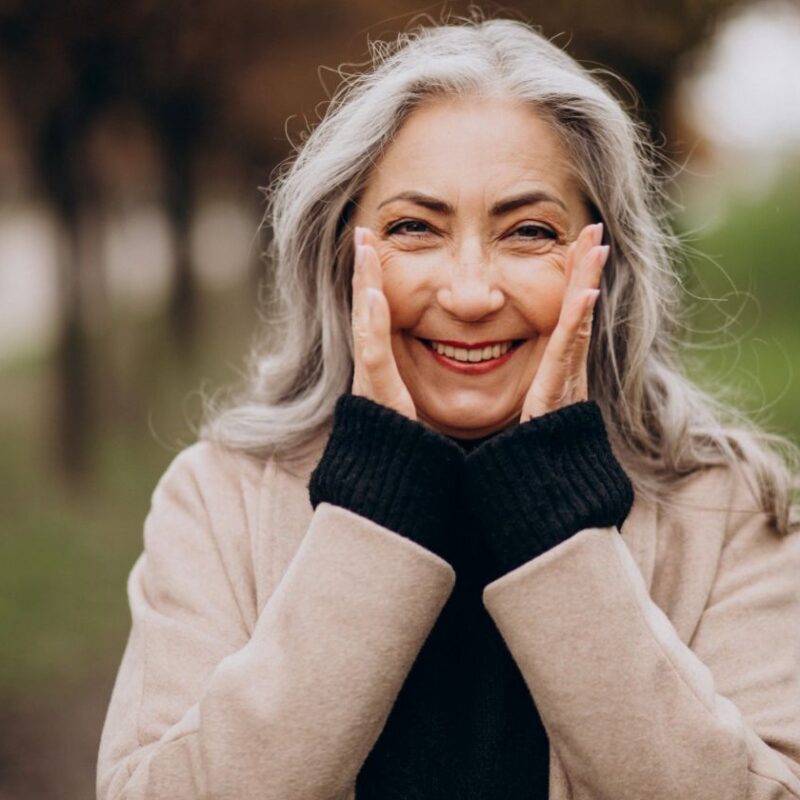 A woman with gray hair smiling with her hands on her cheeks for a blog post titled, "7 Powerful Benefits of Hearing Aids."
